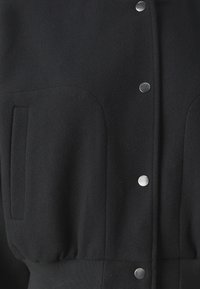 Zwarte knoopjasje met zilveren drukknoopsluitingen, voorzien van grote voorzakken en een gathered taille. Gemaakt van zachte, gestructureerde stof.