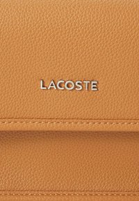 Tan leather wallet with a textured finish, featuring a silver "LACOSTE" logo and tonal stitching for a sleek appearance.