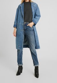 Blauer Jeans-Langmantel mit lockerer Passform, Knopfschluss und großen Taschen. Getragen über einem grauen Oberteil und kombiniert mit ausgewaschenen Jeans und schwarzen Stiefeletten.