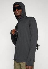 Dark gray long-sleeve hooded shirt with a textured fabric, featuring thumbholes and a small logo, worn over an olive green outfit.