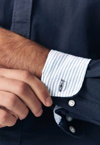 Marineblauw shirt met een boord in blauw en witte strepen. Voorzien van contrasterende stiksels en een knoop. Het boord heeft een klein logodetail vlak bij de opening.