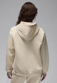 Persona con lunghi capelli scuri che indossa un grande hoodie color crema e pantaloni coordinati, di spalle su uno sfondo grigio uniforme.