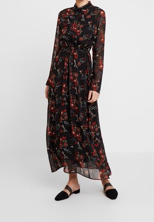 Robe longue noire avec des motifs floraux rouges, en tissu transparent, à manches longues, avec lien à la taille et une silhouette fluide. Associée à des chaussures plates noires.