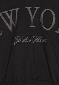Svart sweatshirt med stora, texturerade bokstäver "NEW YORK" och frasen "United States" i kursiv stil, med ett ribbat nederstycke.