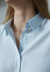 Lyseblå skjorte med knaplukning og struktureret krave, lavet af et glat stof. Har perlemorsknapper og en pæn finish.
