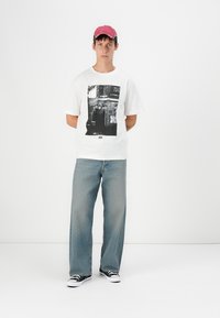 Hvid kortærmet t-shirt med et sort-hvidt grafisk print, parret med lyseblå brede jeans og sorte sneakers. Rød kasket.