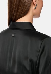 Zwarte satijnen blouse met een gestructureerde kraag en een slank ontwerp, met een knoopdetail aan de achterkant. De gladde textuur versterkt de elegantie.