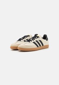 Cream-colored sneakers with black stripes, black accents, and a gum rubber sole. Features a smooth leather texture and lace-up design.