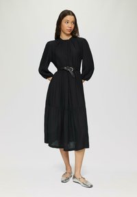 Schwarzes Midikleid mit gerafftem Detail am Ausschnitt, langen Ärmeln und einem gestuften Rock; kombiniert mit silbernen Ballerinas und einem schwarzen Gürtel.