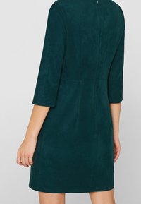 Femme portant une robe verte foncée, midi, à manches trois-quarts, vue de dos et de la taille vers le bas sur fond uni.