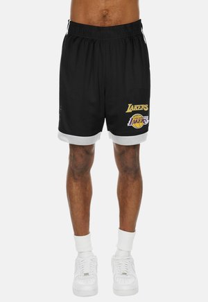 Eine Person trägt schwarze Los Angeles Lakers Basketballshorts mit weißem Rand, weiße Socken und weiße Sneakers und steht vor einem einfarbigen Hintergrund.