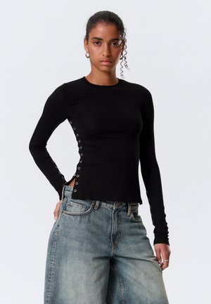 Νεαρή γυναίκα με σγουρά μαλλιά, ντυμένη με μαύρο τοπ μακρυμάνικο με μεταλλικές τρύπες στις πλευρές και ανοιχτό μπλε τζιν παντελόνι φαρδιάς γραμμής.