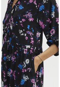 Robe florale en tissu noir avec des motifs vibrants violets, bleus et blancs. Design évasé avec des poches et des manches retroussées.