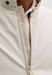 Μπεζ μπουφάν με ψηλό γιακά, φερμουάρ στο μπροστινό μέρος με διακοσμητικές χάντρες και το λογότυπο "JACK & JONES PREMIUM" κατά μήκος του φερμουάρ.