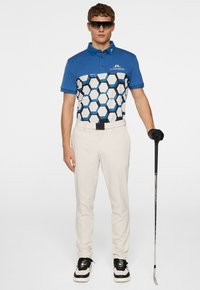 Kortärmad blå polo med hexagonmönster i svart, vitt och blått, tillsammans med beige byxor och svarta sneakers, hållande en golfklubba.