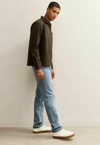 Chaqueta de botonadura oscura verde, jeans azul claro, zapatillas blancas con suelas de goma, tela texturizada y un corte casual. El modelo posa frente a un fondo liso.