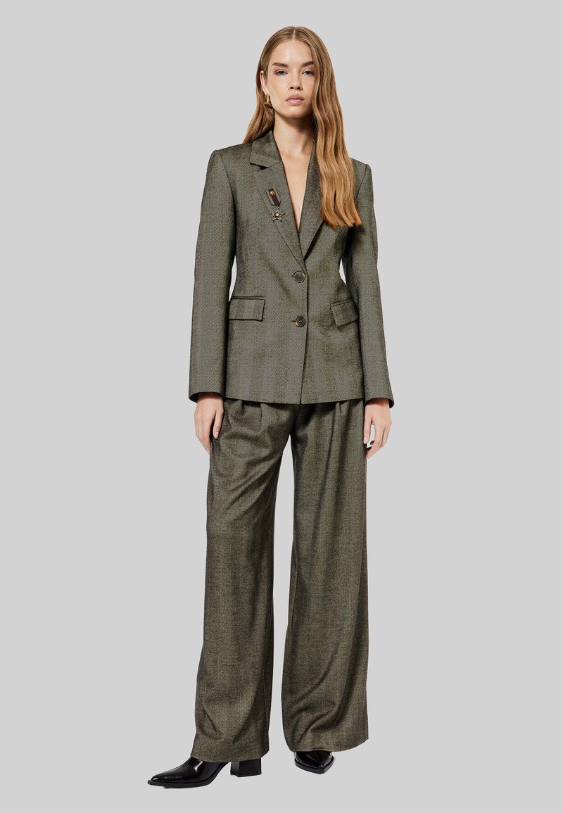 Olijfgroene getailleerde blazer met puntige revers, twee knopen en zijzakken; wijde broek in bijpassende stof; gedragen met zwarte enkellaarsjes.