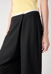 Un pantalon noir présente un design structuré avec des plis à l'avant, une taille cintrée et une texture de tissu lisse.