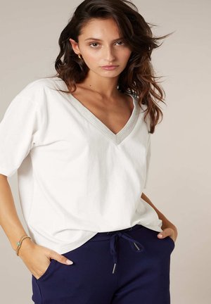 Jeune femme aux cheveux bruns ondulés, portant un t-shirt blanc à col en V et un pantalon marine à cordon, posant les mains dans les poches sur un fond neutre.