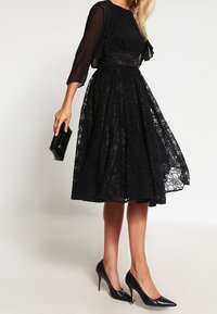 Czarna koronkowa sukienka z dopasowanym gorsetem i zwiewną spódnicą, z rękawami do połowy przedramienia i teksturowanym paskiem w talii. W połączeniu z czarnymi szpilkami.