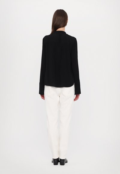 Black long-sleeve blouse with a pleated back detail, paired with white trousers and black shoes. Smooth fabric, tailored fit.