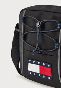 Borsa in tessuto nero con superficie testurizzata, dettaglio in corda elastica nella parte anteriore e patch del logo "TOMMY JEANS" multicolore.