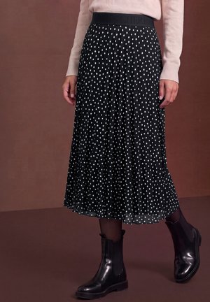 Un mannequin porte une jupe midi plissée noire à pois blancs, un pull rose pâle, des collants noirs et des bottines noires.