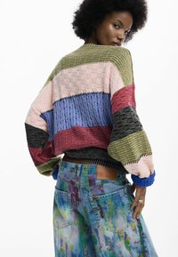 Suéter de punto multicolor con patrón texturizado, que presenta franjas verdes, rosas, azules, rojas y negras; combinado con pantalones cortos de mezclilla salpicados de pintura.