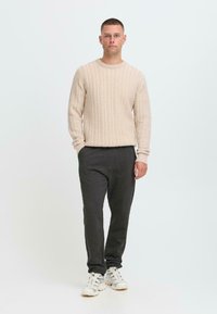 Ung mann som står med hendene delvis i lommene, iført lys beige kabelstrikket genser, mørke grå bukser og hvite sneakers, mot en ensfarget bakgrunn.