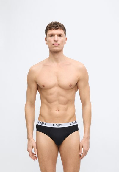 Lihaseline mees seisab näoga ettepoole, seljas mustad püksikud valge vöökohaga, millel on logo, ühevärvilisel heledal taustal.