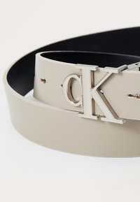 Beige leren riem met een glanzende zilveren 'ck' gesp. De riem heeft een gladde textuur en is ontworpen met meerdere verstelgaten.