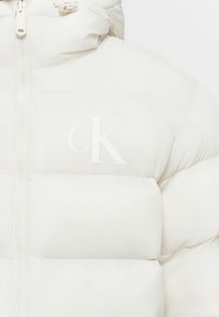 Veste doudoune blanche avec fermeture éclair sur le devant et logo "cK" imprimé en léger sur la poitrine, dotée d'un col haut et d'un matelassage rembourré.