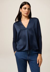 Navyblaues Oberteil mit V-Ausschnitt und langen Ärmeln; aus glattem Stoff gefertigt. Hat eine lockere Passform und einen leichten Fall an der Vorderseite. Kombiniert mit Denim.