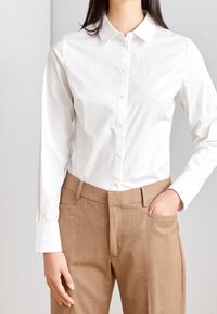 Chemise blanche à boutons avec col classique, associée à un pantalon beige sur mesure avec une poche avant et une texture lisse.