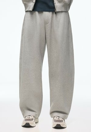 Personne portant un pantalon de survêtement gris ample, des baskets blanches et grises, et une partie d'une veste zippée grise visible sur un t-shirt foncé.