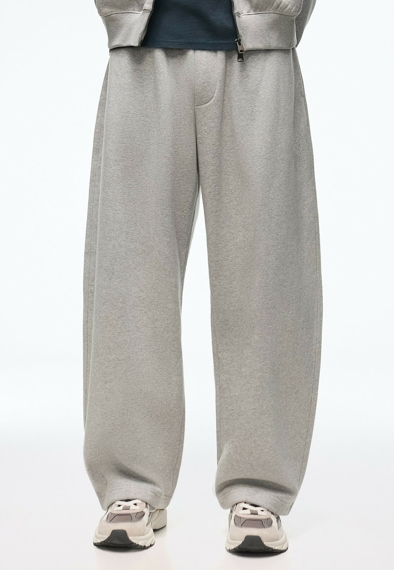 Persoon draagt losse grijze joggingbroek, witte en grijze sneakers, en een deel van een grijze zip-up jas zichtbaar over een donker shirt.
