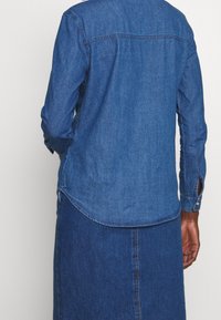 Denim shirt in deep blue with a curved hem, buttoned cuffs, and a classic collar. Features a back yoke and soft, textured fabric.