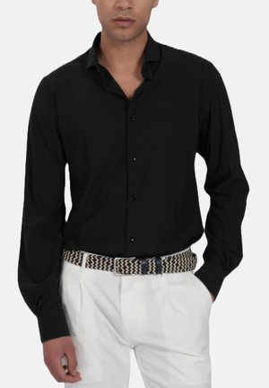 Uomo che indossa una camicia nera a maniche lunghe con bottoni, pantaloni bianchi e cintura intrecciata con motivo bianco e nero, mani nelle tasche, su sfondo semplice.