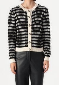 Kobieta ubrana w czarną i beżową swetrową kardigan z dużymi guzikami, w połączeniu z czarnymi skórzanymi spodniami, stojąca na tle białym.