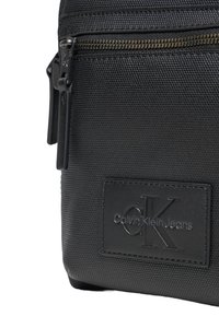 Bolso de hombro negro de nailon con una superficie texturizada, cierre de cremallera y una etiqueta con el logotipo que presenta letras en relieve; forma compacta y rectangular.