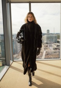 Naine pikkade tumedate karvase mantliga, mustade kinnaste ja saabastega kõndimas siseruumides, hoides suurt musta käekotti, kõrgete akende kaudu on näha linnasiluetti.