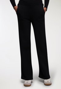 Pantaloni di felpa neri a gamba larga con fascia elastica in vita e tasche laterali, realizzati in un tessuto morbido e liscio. Indossati con scarpe casual.
