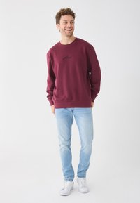 Sweat-shirt bordeaux avec col rond et logo noir, associé à un jean bleu clair et des baskets blanches, sur un fond uni.