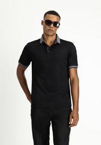 Schwarzes Poloshirt mit kurzen Ärmeln, grauem Kragen und Akzenten an den Bündchen sowie einer Knopfleiste. Das Model trägt schwarze Jeans und Sonnenbrillen.