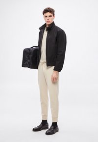 Czarna lekka kurtka, beżowa koszula z kołnierzykiem, beżowe spodnie dresowe i czarne mokasyny. Model trzyma małą czarną torbę. Prosty, nowoczesny design.