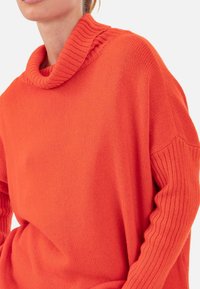 Pull orange en maille avec un col châle côtelé et des manches texturées. Le tissu semble doux et confortable avec une coupe décontractée.