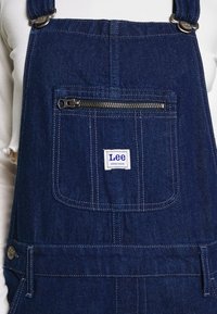 Ogrodniczki dżinsowe w ciemnoniebieskim kolorze, z przednią kieszenią na zamek oraz białą naszywką z logo. Widoczne metalowe zatrzaski i detale w kontrastowym przeszyciu.