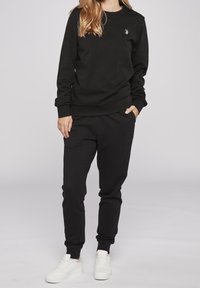 Černá mikina s dlouhými rukávy a žebrovanými manžetami, v kombinaci s černými tepláky. Oblečení doplňují bílé tenisky. Vzhled umocňuje decentní detail loga.