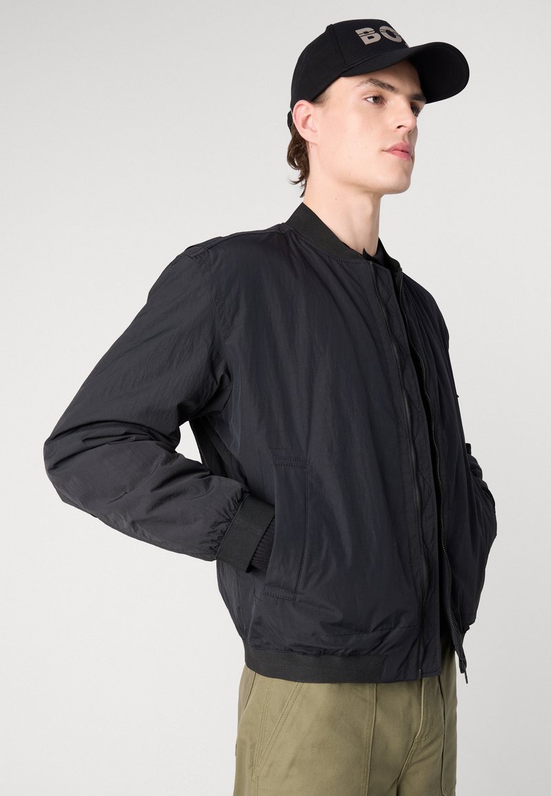 Czarna nylonowa kurtka bomberka o gładkiej fakturze, z prążkowanymi mankietami i dołem, z suwakiem z przodu oraz bocznymi kieszeniami, noszona na zielonych spodniach.