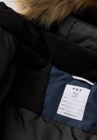 Sort dunjakke indvendig med blødt mørkegrå fleecefor, et blåt stofpanel og en hvid label med størrelse og plejeanvisninger.